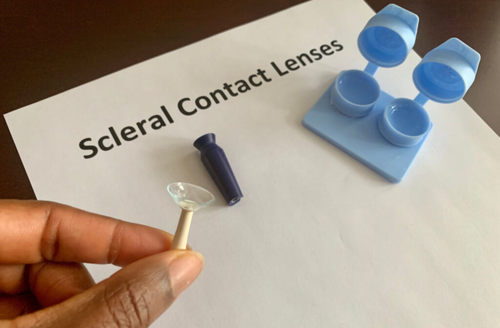 A hand holds an insertion tool for contact lenses, with a scleral lens delicately secured at the tip, with a container for contact lenses and a removal tool in the background.